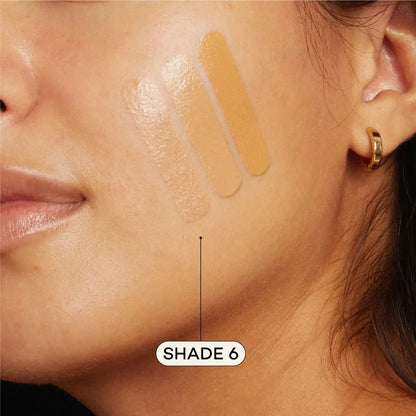 A close-up of a woman’s face with three Axiology Fungdation Mushroom Foundation swatches in different shades on her cheek. An arrow labels the darkest as SHADE 6. She wears a small gold hoop earring.