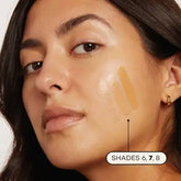 A woman with medium skin tone wears Axiology Fungdation Mushroom Foundation in shades 6, 7, and 8 swatched on her cheek. She has dark hair, minimal makeup, and small gold hoop earrings.