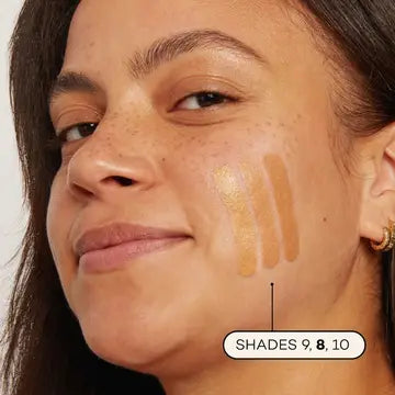 A woman with medium skin tone shows three Axiology Fungdation Mushroom Foundation swatches—shades 9, 8, and 10—on her cheek to compare colors. She smiles gently while facing the camera.