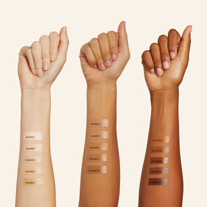 Three raised forearms with different skin tones display twelve swatches of Axiology Fungdation Mushroom Foundation, labeled by shade number and showing a spectrum of foundation colors from light to dark on each arm.