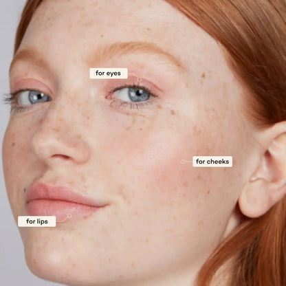 A close-up of a woman with fair skin, red hair, and blue eyes features Axiology 3-in-1 Balmie Crayon Singles applied on eyelids, cheekbones, and lips—highlighting plastic-free makeup versatility. 