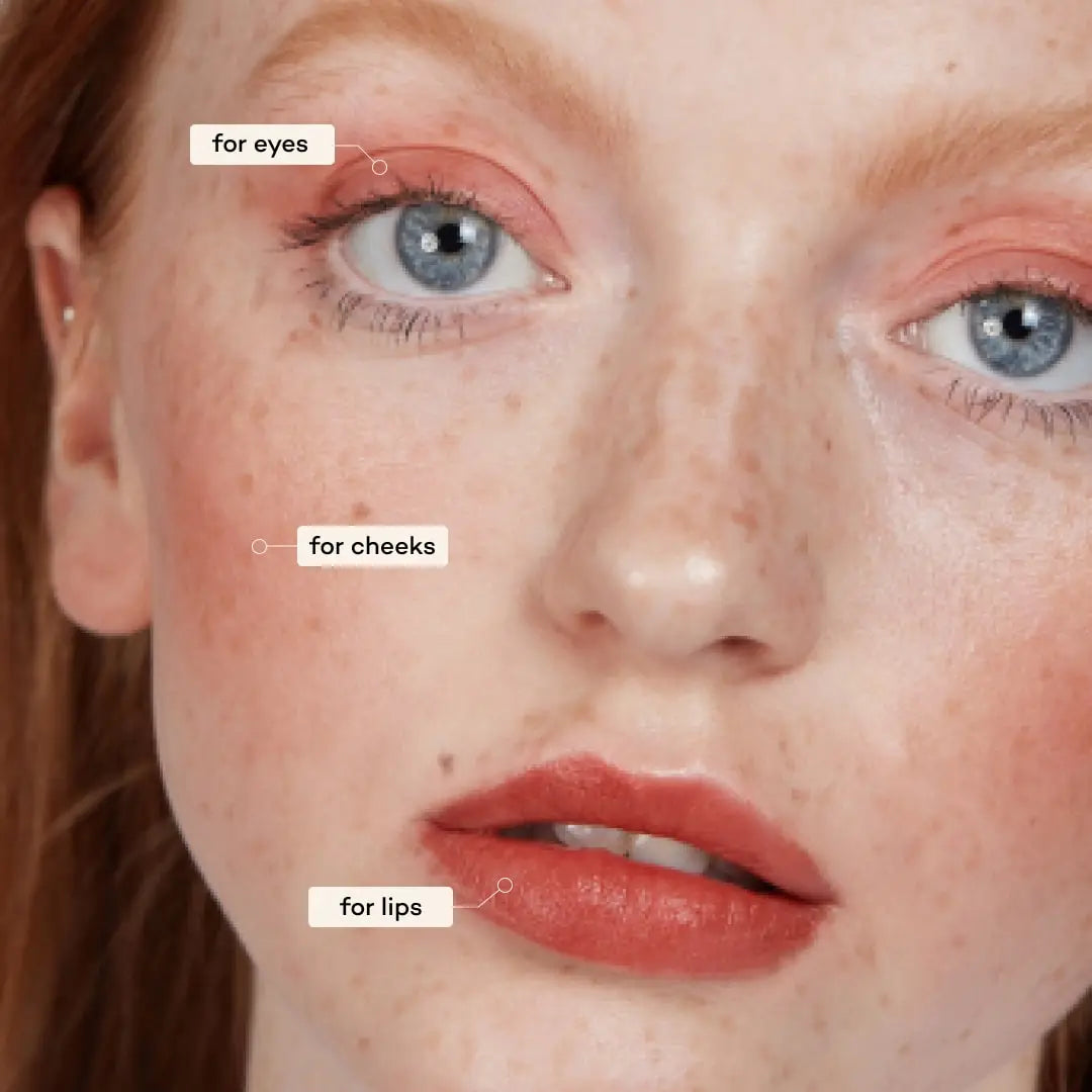 Close-up of a fair-skinned, red-haired person wearing peach matte color on eyes, cheeks, and lips—each highlighted as "for eyes," "for cheeks," and "for lips"—created with Axiology 3-in-1 Balmie Crayon Singles. 