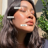 A person with long dark hair faces sunlight outdoors, eyes closed, wearing glowing eye, cheek, and lip makeup created with Axiology 3-in-1 Balmie Crayon Singles in plastic-free packaging. Green foliage and a beige wall are behind. 