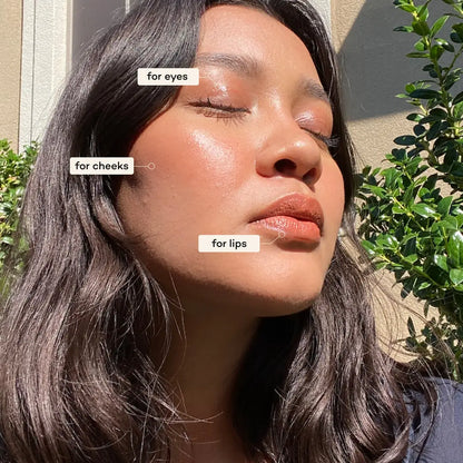 A person with long dark hair faces sunlight outdoors, eyes closed, wearing glowing eye, cheek, and lip makeup created with Axiology 3-in-1 Balmie Crayon Singles in plastic-free packaging. Green foliage and a beige wall are behind. 