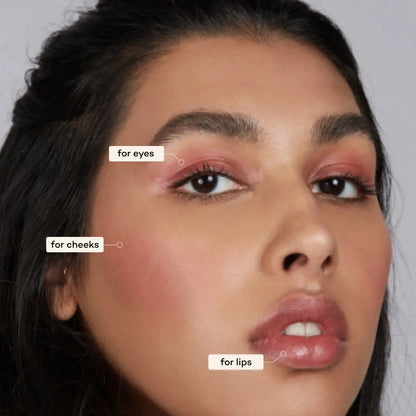 A close-up shows pink makeup on a woman’s eyelids, cheeks, and lips, applied with Axiology 3-in-1 Balmie Crayon Singles. Labels point to each area, reading “for eyes,” “for cheeks,” and “for lips.”. 