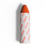 Axiology’s 3-in-1 Balmie Crayon Single in a short, orange stick partially wrapped in white paper printed with ”rosé” in pink—a perfect plastic-free choice for eco-conscious makeup lovers. 