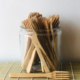 A glass jar holds Bamboo Switch Individual Bamboo Utensils with one fork in front on a bamboo mat. The natural wood-colored forks feature a simple, eco-friendly design, making them a sustainable utensil choice.