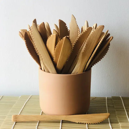 A tan ceramic cup holds several Bamboo Switch Individual Bamboo Utensils on a bamboo mat, providing an eco-friendly option, with one sustainable utensil placed in front of the cup.