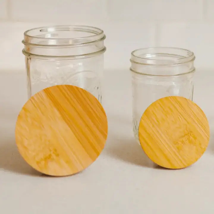 Two empty glass jars of different sizes stand on a white surface, each with a Zefiro Standard or Wide Mouth Solid Bamboo Jar lid resting in front. The background features a light-colored tiled wall.