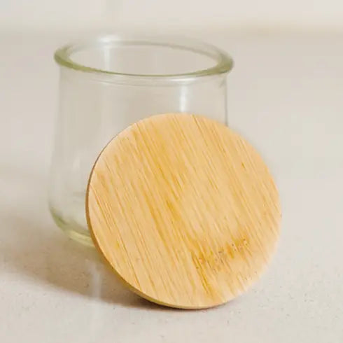 A small, empty glass jar with a round, flat Yogurt Jar Bamboo Lid by Bamboo Switch resting in front—ideal for anyone wanting reusable bamboo jar lids.