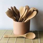 A beige container holds several Bamboo Switch Individual Bamboo Utensils—sustainable wooden spoons in assorted shapes—while one rests on a bamboo mat, highlighting this eco-friendly option against a plain white wall background.