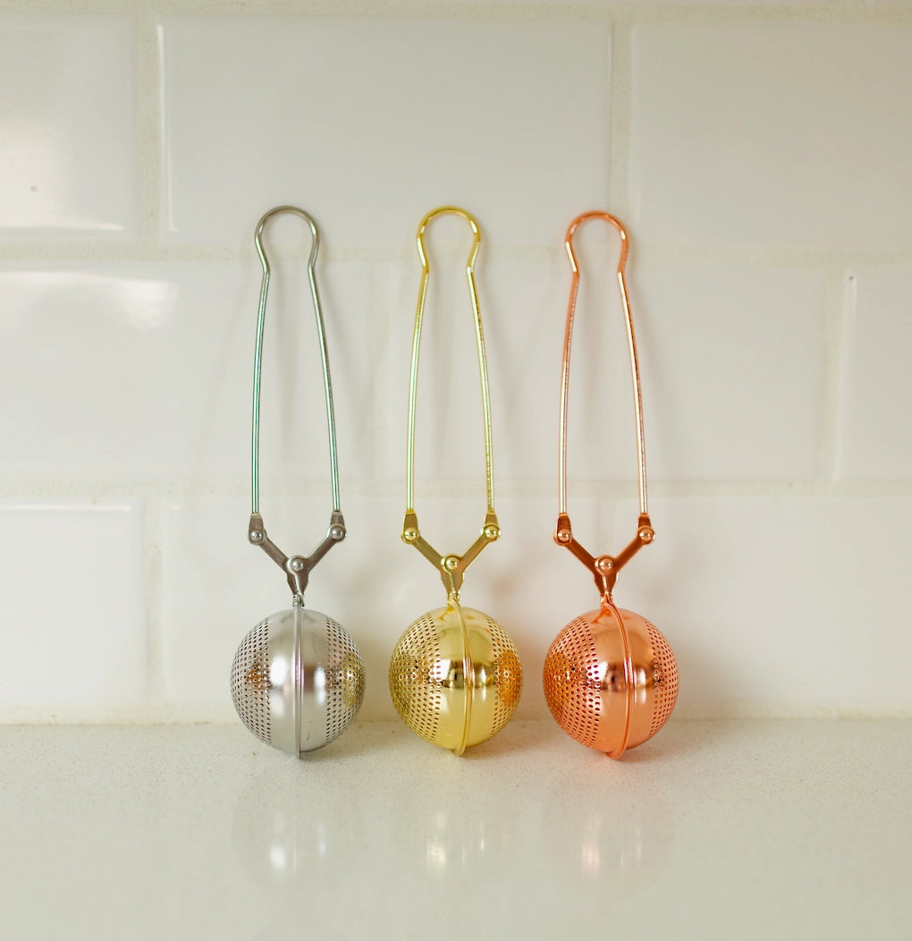 tea strainers lined up against white tile in steel, gold, and rose gold