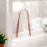 rose gold tongue scraper propped up against white bathroom tile