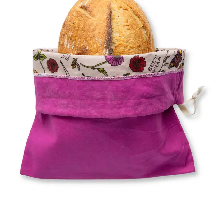 Purple reusable bread bag with floral trim holding a loaf of bread on a white background