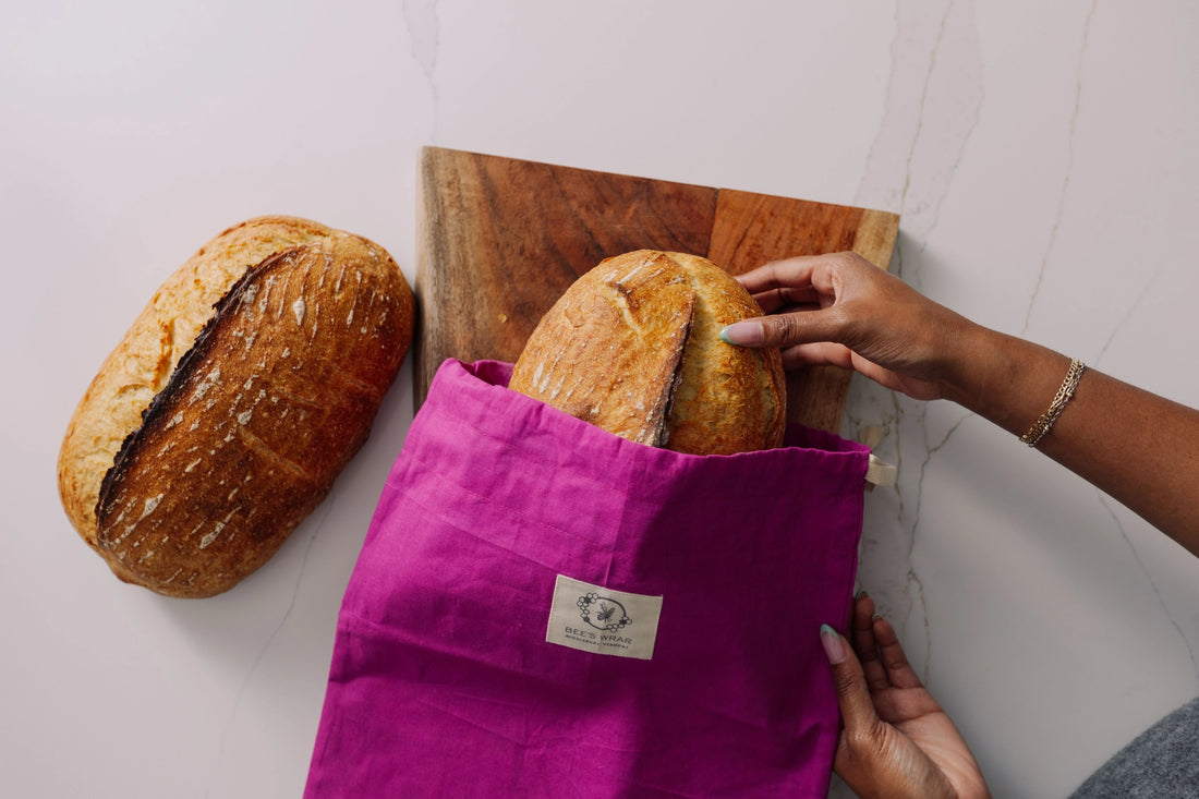 Person placing bread into a purple reusable bread bag on a white surface with a wooden cutting board.