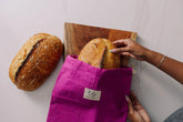 Person placing bread into a purple reusable bread bag on a white surface with a wooden cutting board.