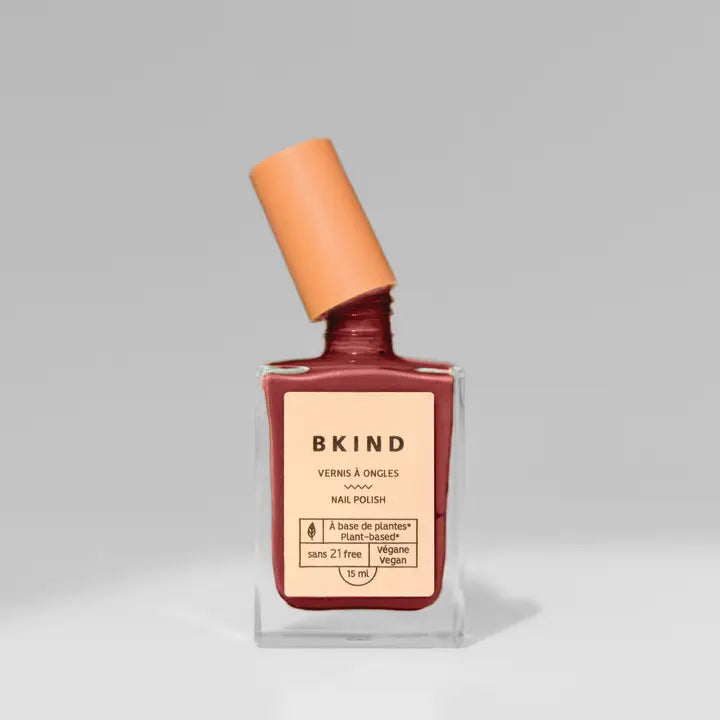 A bottle of BKind Nail Polish with a peach cap, slightly tilted, displays its plant-based, 21-free formula in a reddish-brown shade against a light gray background.