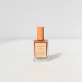 A bottle of BKind Nail Polish by BKind, featuring a peach cap and label and filled with muted pink polish, rests on a plain white surface with a soft shadow.