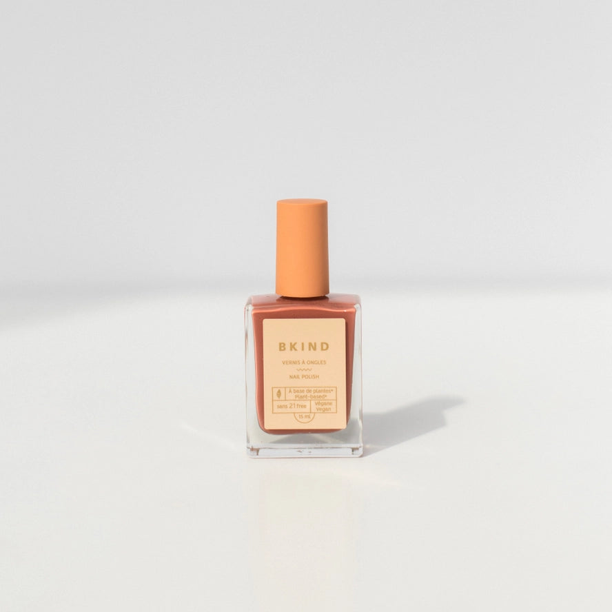 A bottle of BKind Nail Polish by BKind, featuring a peach cap and label and filled with muted pink polish, rests on a plain white surface with a soft shadow.
