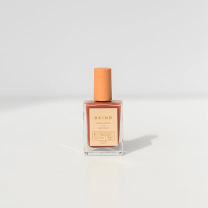 A bottle of BKind Nail Polish by BKind, featuring a peach cap and label and filled with muted pink polish, rests on a plain white surface with a soft shadow.