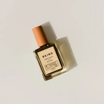 A square glass bottle of BKind Nail Polish by BKind with a rose gold cap sits on a light surface, casting a gentle shadow. The front label is clearly visible.