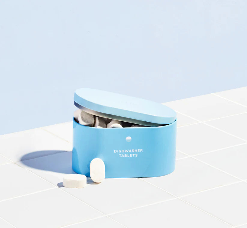 A light blue Blueland Dishwasher Tablets container, labeled Plastic-Free, rests on white tiles with some white tablets spilling out. The pale blue background highlights this zero waste dishwasher detergent.