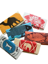 Six colorful rectangular Cement6 Accordion Clutch bags, each artisan made with bold graphic illustrations—a camel, a flower, two types of fish, a tiger, and an elephant—are thoughtfully arranged on a white background.