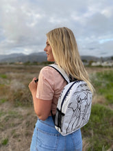 A woman with long blonde hair wears a pale pink t-shirt, blue jeans, and carries the Cement6 Minimal Daypack featuring black elephant illustrations, standing in a grassy field with mountains in the background.