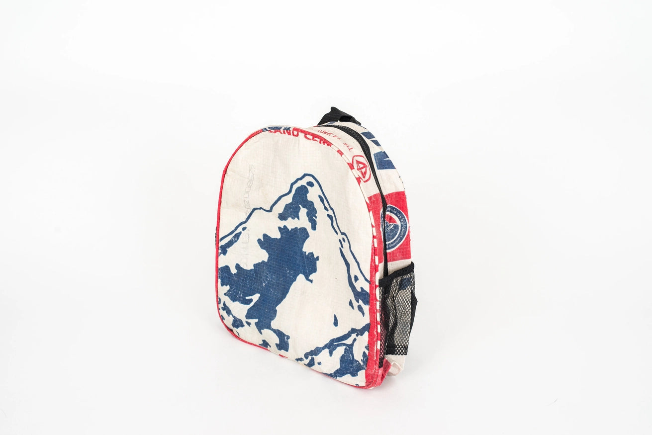 The Cement6 Minimal Daypack is a small, white artisan backpack with red trim, a blue mountain design on the front, and black mesh side pockets, shown standing upright against a plain white background.