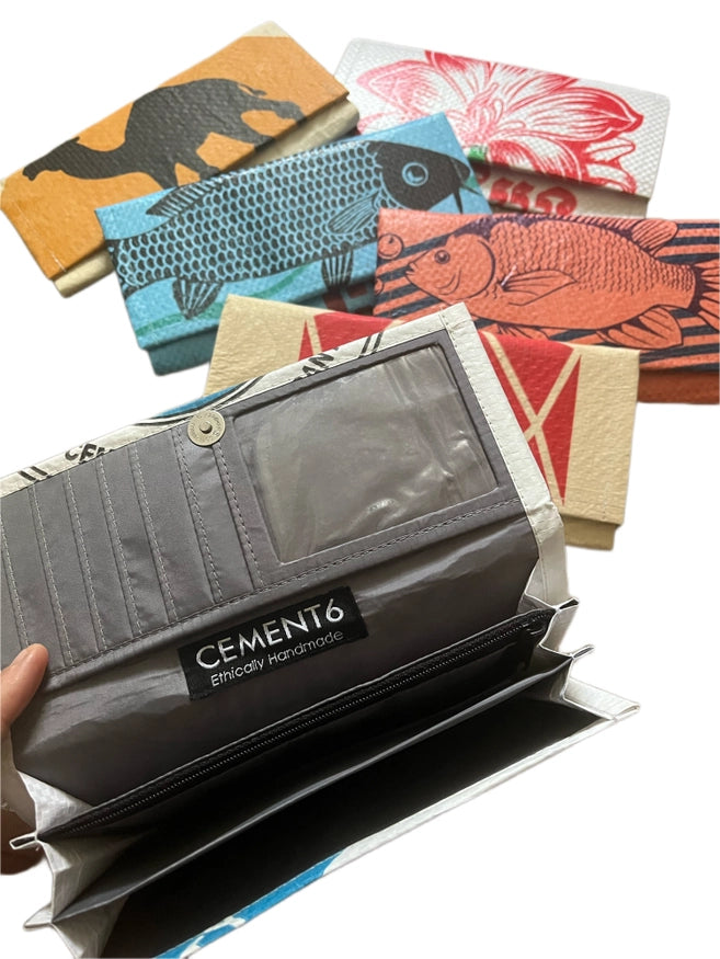 A hand displays an Accordion Clutch by Cement6, featuring multiple compartments. Behind it, five colorful wallets with animal and fish designs highlight the brand&