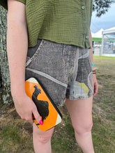 A person outdoors in a green textured shirt and patterned shorts holds the Cement6 Zipped Clutch featuring a black rooster silhouette. An ethical wallet is visible, and their bright pink nail polish adds a pop of color.
