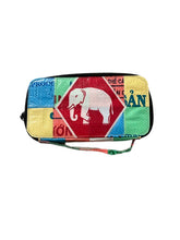 The Cement6 Zipped Clutch is a colorful, rectangular artisan bag with patchwork, a central red diamond and white elephant, green, yellow, and blue blocks. Embrace sustainable style with unique patterns and a handy wrist strap.