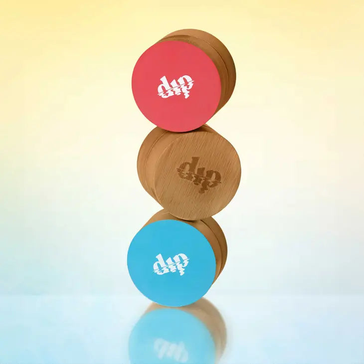 Three Dip Bamboo Travel Cases with Coasters are stacked vertically: the top with a pink lid, the middle plain, and the bottom with a blue lid. Each features a white logo, ideal for stylishly storing shampoo bars. Background: soft yellow-to-blue gradient.