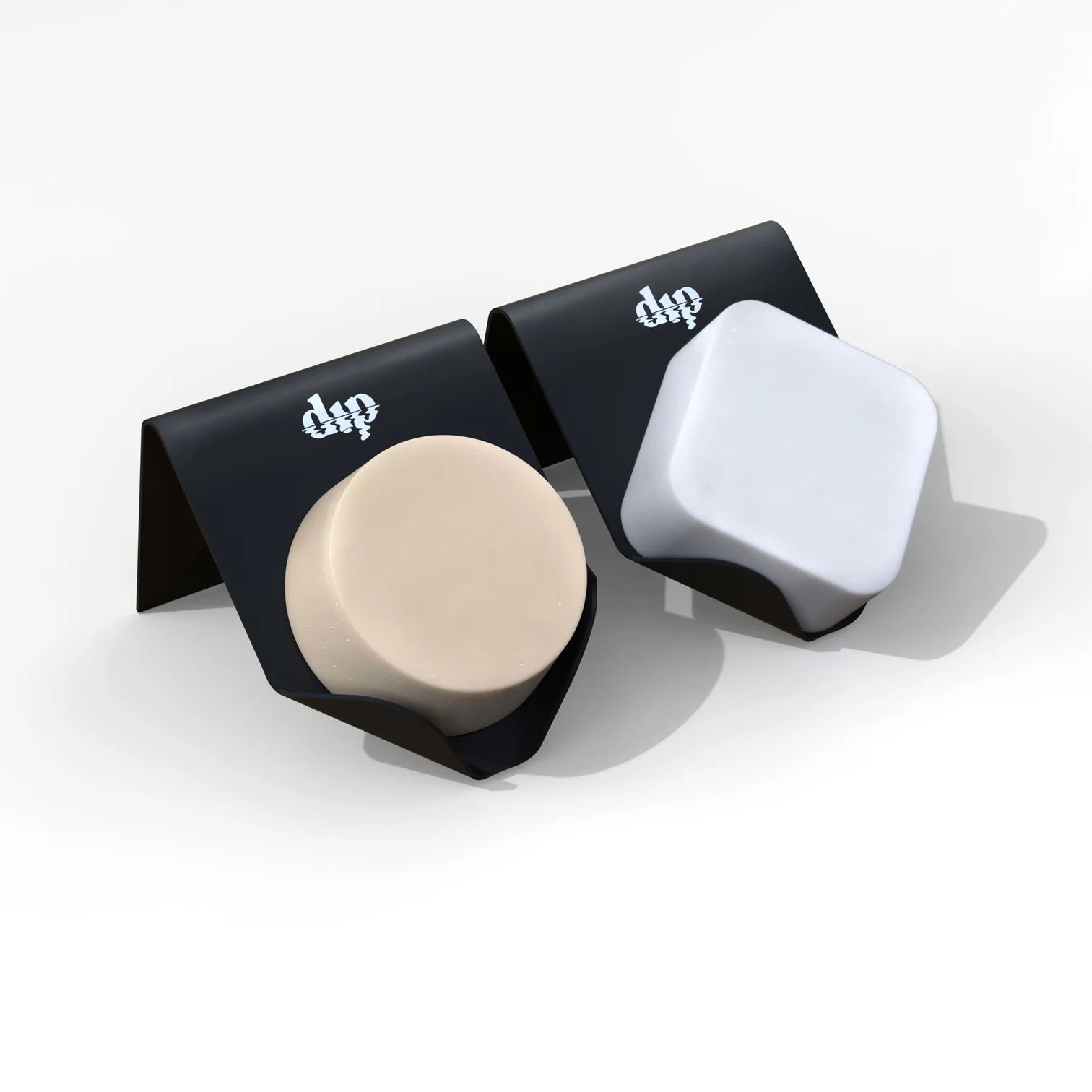 Two black, durable Dip Life Preserver Draining Soap Dishes, each holding a soap bar—one beige and round, the other white and square—are shown on a white background. These eco-friendly holders feature a stylized dp logo on top.