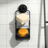 A black Dip & Drip Wall Mount Adapter by Dip, secured with a twist-lock suction cup to a white tiled wall, features two shelves holding a square beige soap above and round orange soap below, both slightly wet.