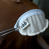 A close-up of the Ebb Handheld Brewer by GDS Cloth Goods—a small, white, organic cotton coffee filter pouch with a scalloped edge and ebb tag—held by metal tongs against a blurred brown shirt background.