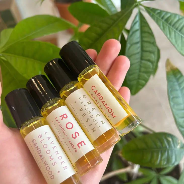 A hand holds four FD Market Perfume Rollers in glass bottles, labeled LAVENDER BLOOMS + SANDALWOOD, ROSE PATCHOULI + AMBER, GREEN PATCHOULI + RESIN, and CARDAMOM SAFFRON. A leafy plant is visible in the background.