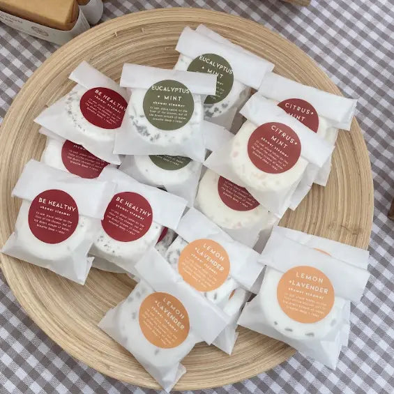 Assorted packets of different scented shower steamers on a wooden plate with a checkered tablecloth background.