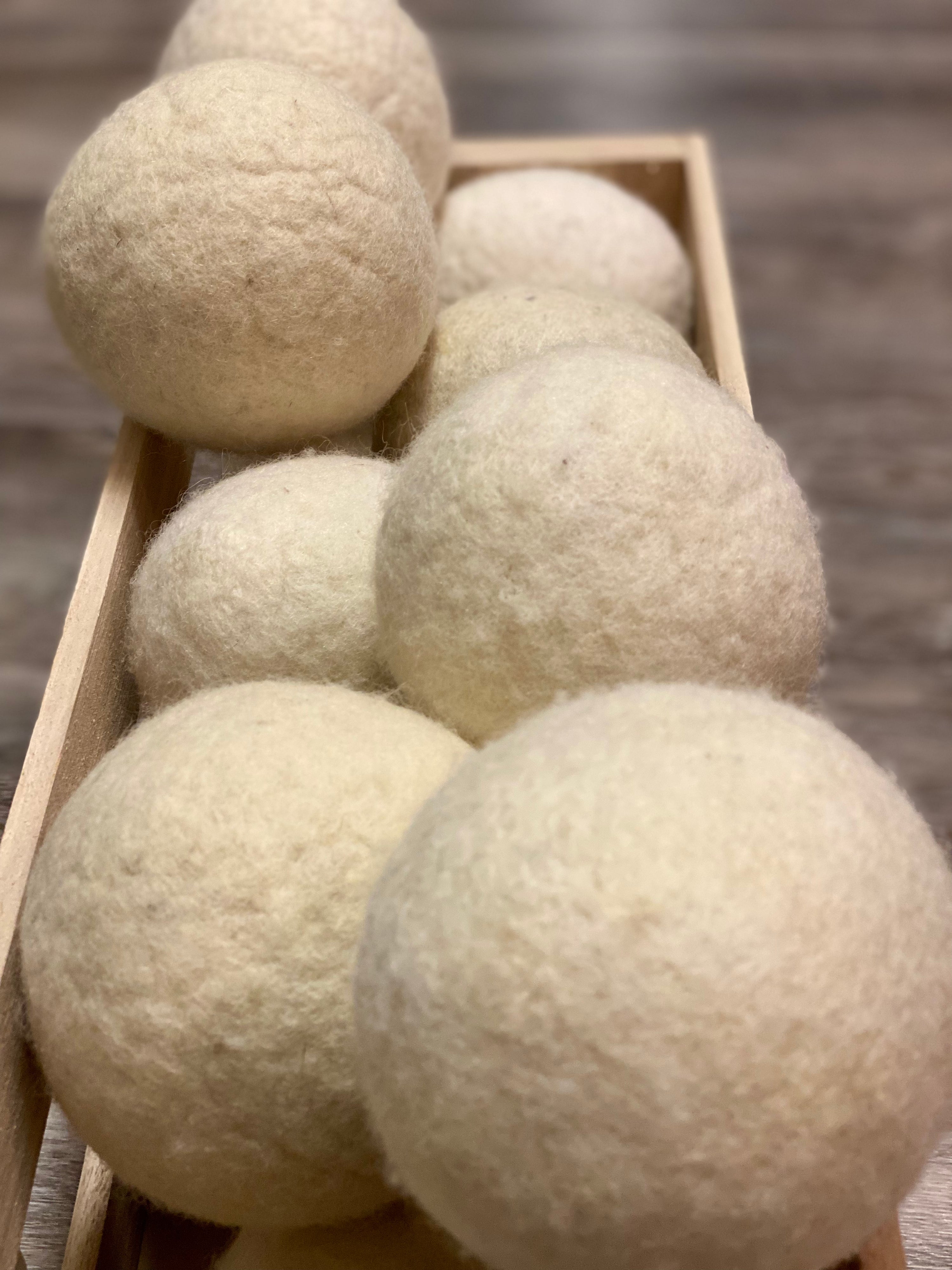 A wooden tray holds several Jumbo Dryer Balls by Green Gifts, large off-white wool balls that serve as an eco-friendly fabric softener. They appear soft and slightly fuzzy, resting on a wood-textured surface.
