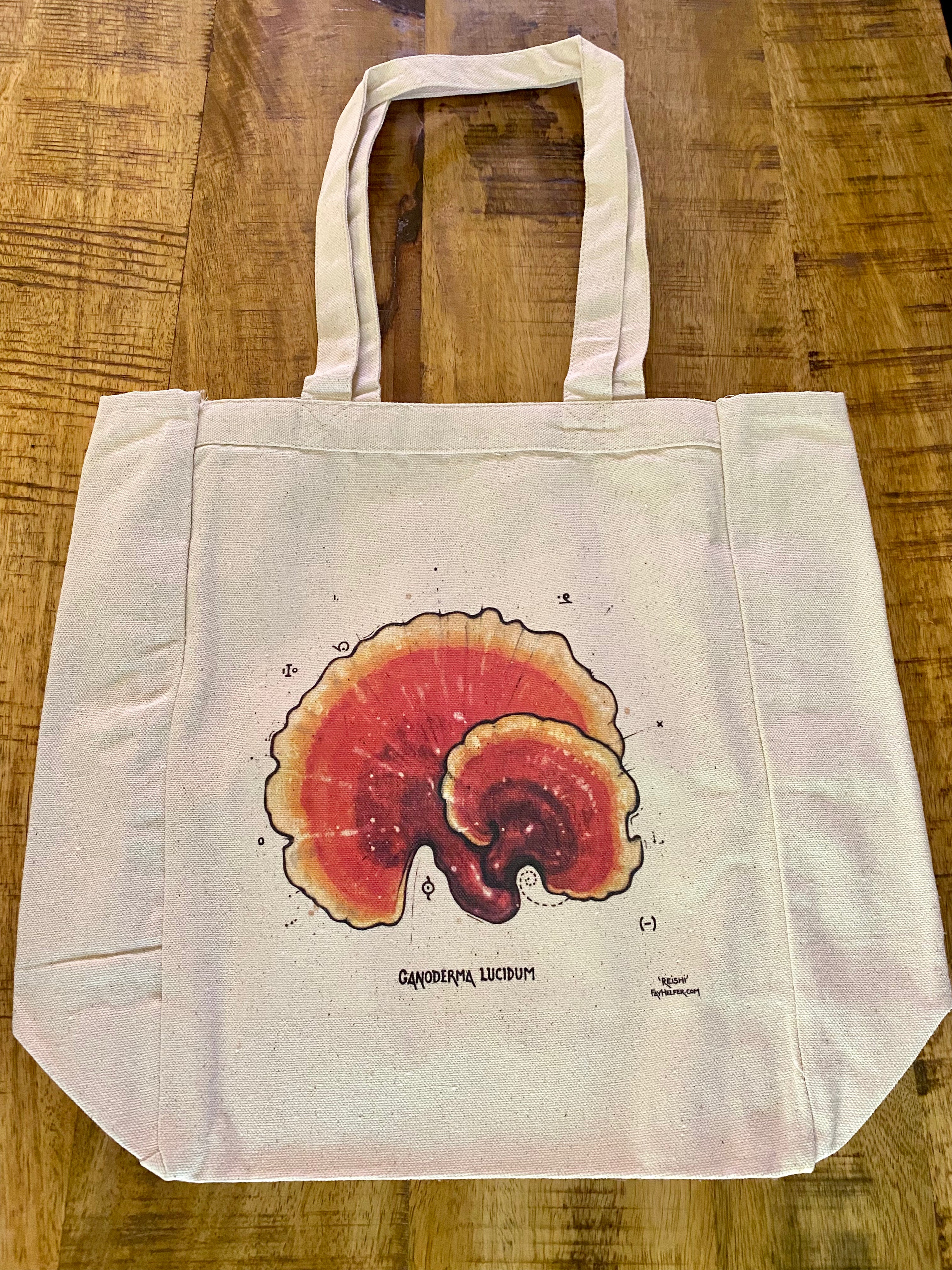 The Fay Helfer Mushroom Tote Bag, made of beige eco cotton, features a reddish-brown Ganoderma lucidum mushroom illustration with handwritten labeling, shown resting on a wooden surface—an inviting nod to medicinal mushrooms.