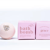 A pale pink Ginger and June Botanical Bath Bomb with dried herbs rests beside two minimalist pink boxes labeled "Bath Bomb" and "Grace" on a clean white background.
