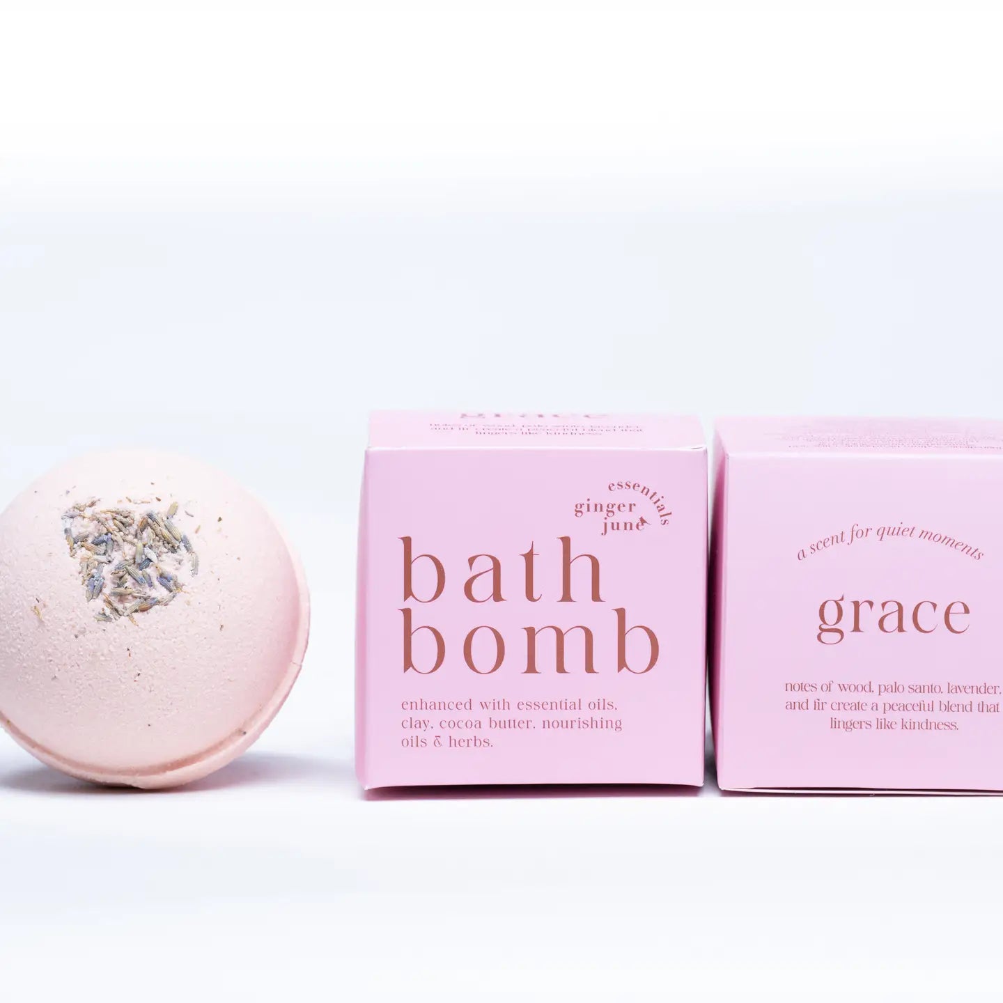 A pale pink Ginger and June Botanical Bath Bomb with dried herbs rests beside two minimalist pink boxes labeled "Bath Bomb" and "Grace" on a clean white background.
