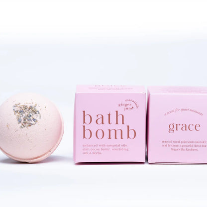 A pale pink Ginger and June Botanical Bath Bomb with dried herbs rests beside two minimalist pink boxes labeled "Bath Bomb" and "Grace" on a clean white background.