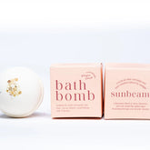 A white Ginger and June Botanical Bath Bomb with a gold leaf accent is displayed next to two pink boxes labeled "Bath Bomb" and "Sunbeam," featuring essential oils, cocoa butter, and a citrus scent blend.