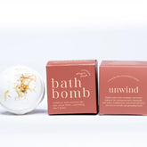 A white Botanical Bath Bomb with dried flower petals from Ginger and June rests beside two pink boxes, featuring essential oils, ginger, juniper, and soothing ingredients for a relaxing bath experience.