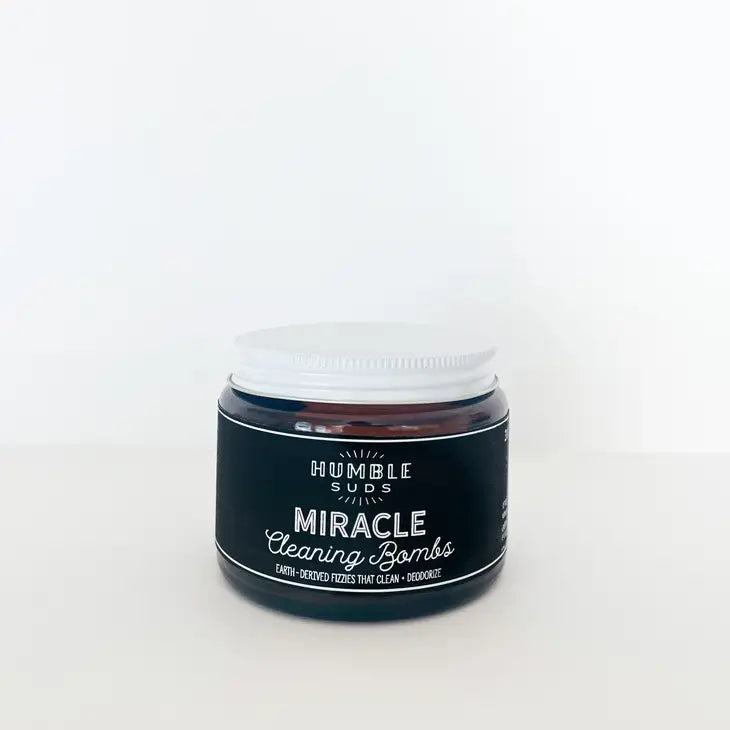 A small round jar of Humble Suds Miracle Cleaning Bombs with a white lid rests on a plain white background, showcasing non-toxic cleaning power and a touch of citrus essential oil.