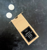 A brown paper bag labeled "Miracle Cleaning Bombs" by Humble Suds sits on a dark surface with two white tablets above it—non-toxic cleaning tablets infused with citrus essential oil for a fresh, powerful clean.