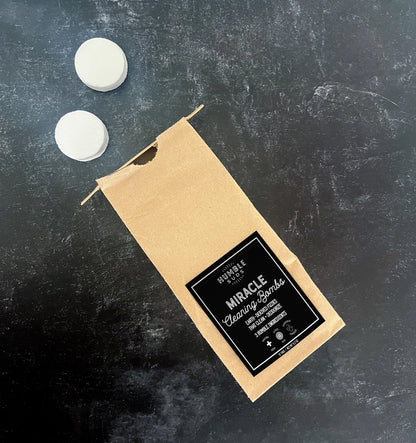 A brown paper bag labeled "Miracle Cleaning Bombs" by Humble Suds sits on a dark surface with two white tablets above it—non-toxic cleaning tablets infused with citrus essential oil for a fresh, powerful clean.