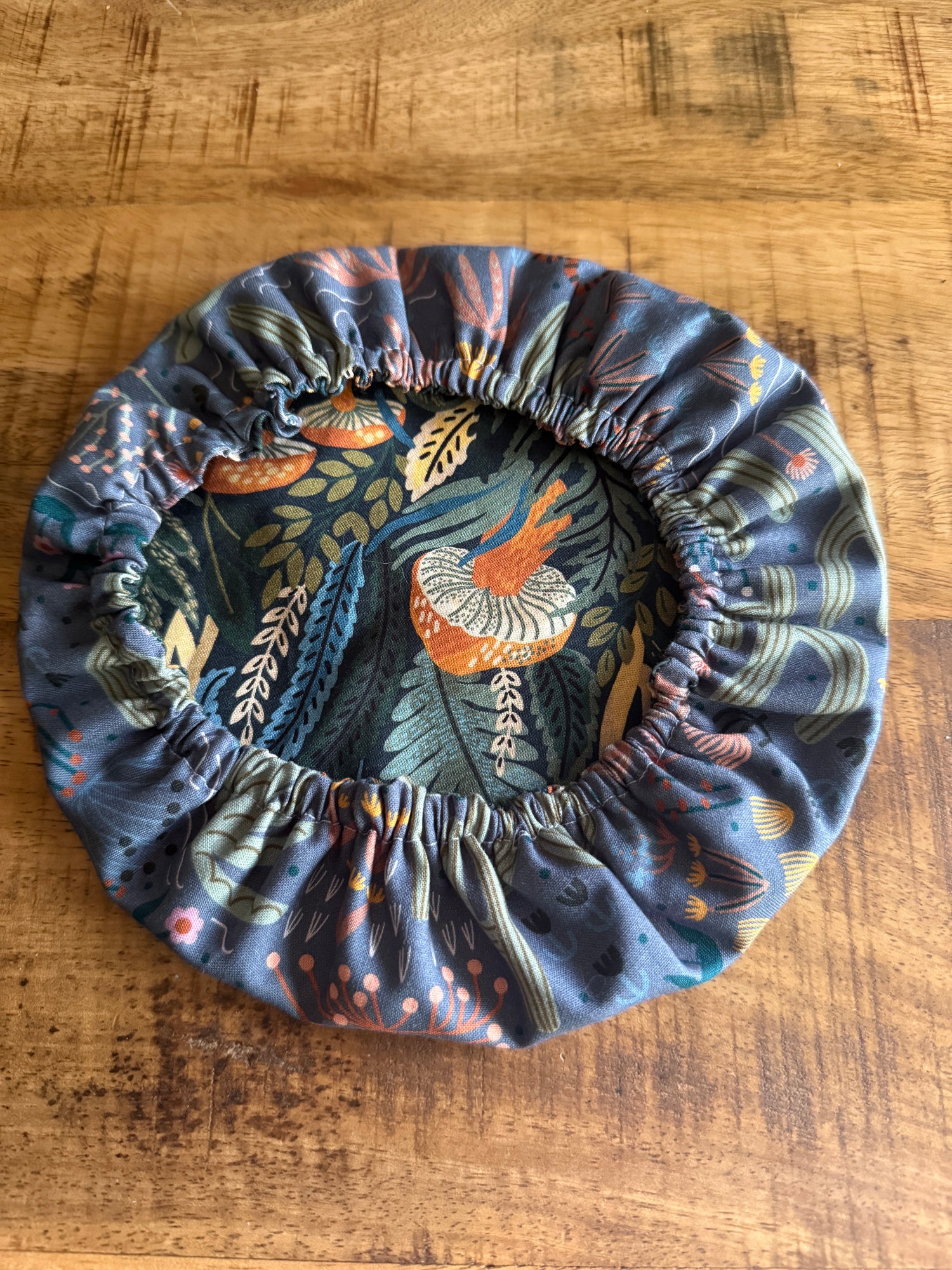 The Sustainable For Good Reusable Reversible Bowl Cover, with elastic edging and a vibrant botanical print of orange, blue, and green leaves plus mushrooms, adds a stylish, plastic-free touch to your kitchen while keeping bowls covered.
