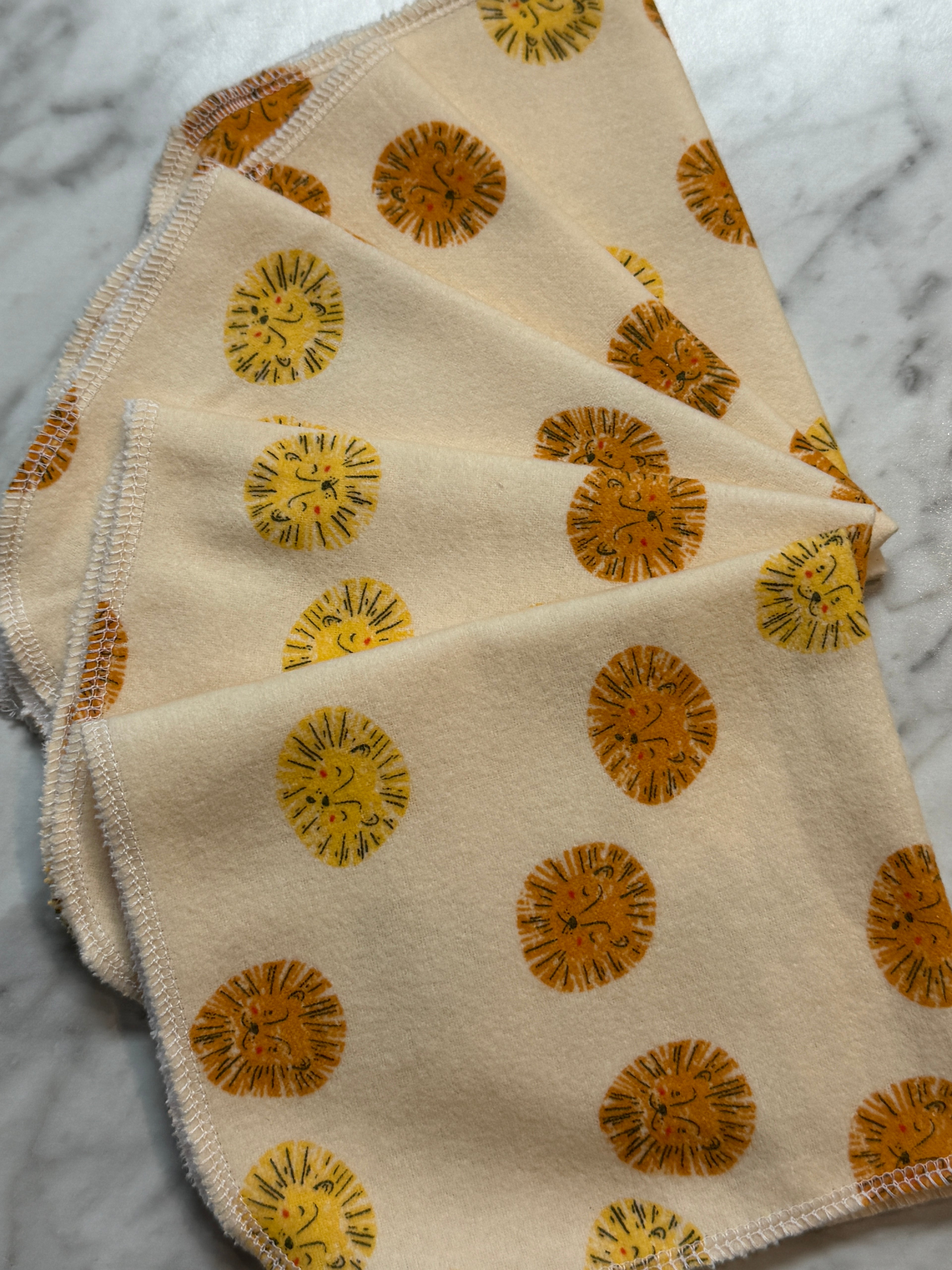 A cream-colored absorbent flannel cloth from Sustainable For Good’s Not Paper Towels Bundles features orange and yellow lion faces, white-stitched edges, and lies on marble—perfect as eco-friendly cleaning or reusable paper towels.